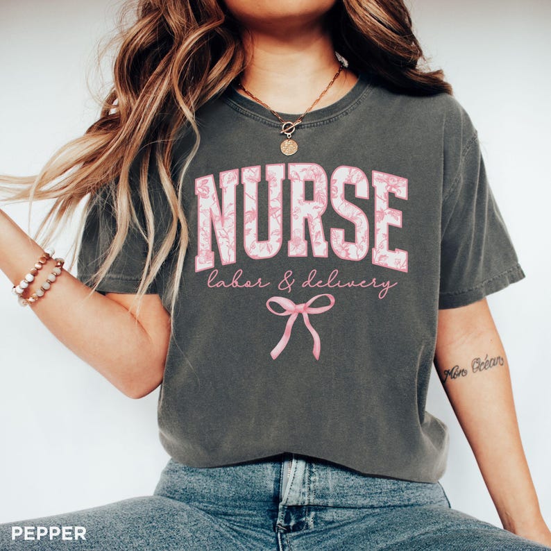 May include: A gray t-shirt with the text "NURSE labor & delivery" in pink and white lettering. The shirt has a pink bow design and a floral pattern.