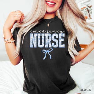 Puede incluir: Camiseta negra con el texto "emergency nurse" en blanco con un patr&oacute;n floral azul. Un lazo azul est&aacute; debajo del texto.