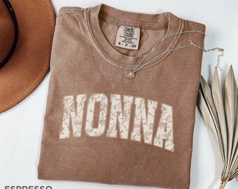 May include: A brown t-shirt with the word "NONNA" printed in white with a floral pattern. The shirt is laid flat on a white surface with a brown hat and a silver necklace.