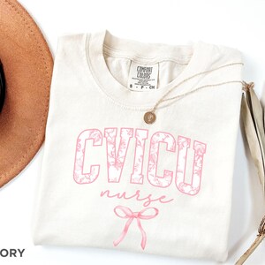 May include: Ivory t-shirt with a pink floral design that says "CVICU nurse" with a pink bow underneath.