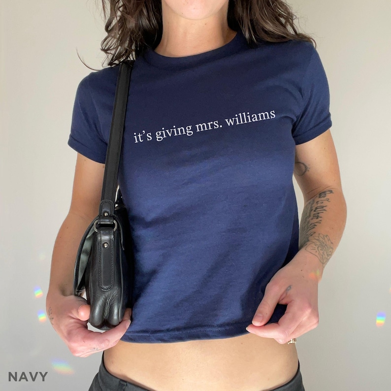 Puede incluir: Camiseta azul marino con el texto "it's giving mrs. williams" en blanco. La camiseta tiene mangas cortas y cuello redondo. Se ve un bolso negro.