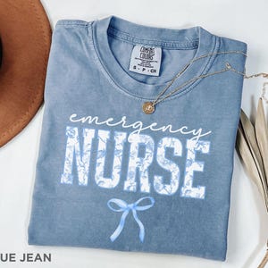 Puede incluir: Una camiseta azul jean con un dise&ntilde;o floral blanco y el texto "emergency nurse" en blanco con un lazo azul debajo. La camiseta est&aacute; hecha por Comfort Colors.