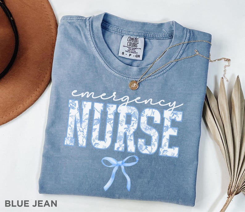Puede incluir: Una camiseta azul jean con un dise&ntilde;o floral blanco y el texto "emergency nurse" en blanco con un lazo azul debajo. La camiseta est&aacute; hecha por Comfort Colors.