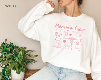 May include: White crewneck sweatshirt with pink lettering that says "Mammo Crew" and pink illustrations of medical symbols, including a stethoscope, a heart, and a breast cancer awareness ribbon.