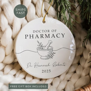 May include: White ceramic ornament with gold string, featuring the text "DOCTOR OF PHARMACY" above a mortar and pestle illustration. Below, it reads "Dr. Hannah Roberts 2025". The ornament is set against a textured white background.