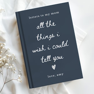 May include: A navy blue journal with white script that reads "letters to my mom, all the things I wish I could tell you, love, Amy." The journal is on a white surface with white flowers.