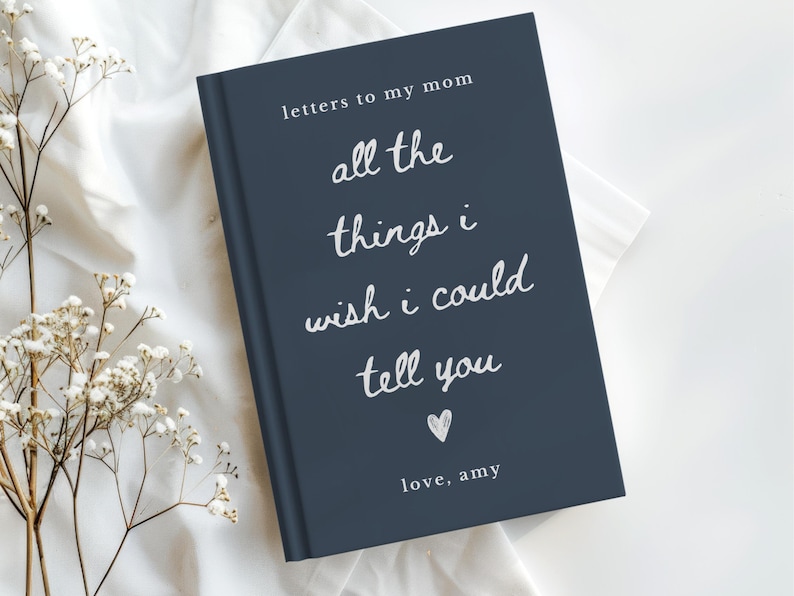 May include: A navy blue journal with white script that reads "letters to my mom, all the things I wish I could tell you, love, Amy." The journal is on a white surface with white flowers.