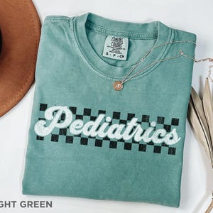 May include: Light green t-shirt with a white checkered pattern and the text "Pediatrics" in a retro font.