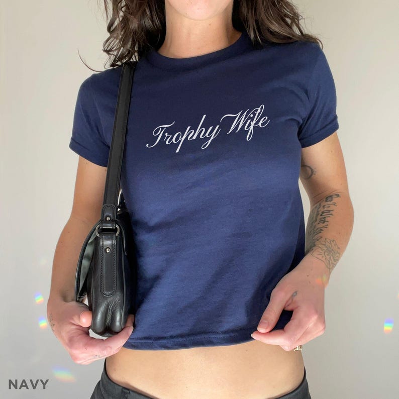 May include: Navy blue crop top with the text "Trophy Wife" in white script. The shirt has short sleeves and a crew neck. A black shoulder bag is visible.