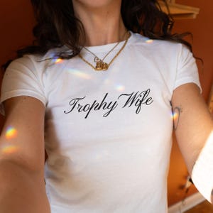 May include: White t-shirt with the words "Trophy Wife" in black script. The shirt has short sleeves and a crew neck. A gold necklace is visible at the neckline. The background is a warm, orange-toned colour.