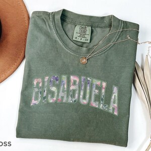 May include: Green t-shirt with the word "BISABUELA" in floral print letters. The shirt is made of a soft, comfortable material.