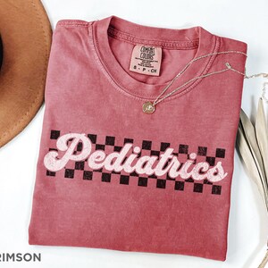 May include: A crimson red t-shirt with a white checkered design and the text "Pediatrics" in a retro font.