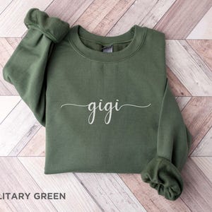 May include: A military green sweatshirt with the word "gigi" written in white cursive script.