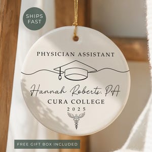 May include: A round, white ceramic ornament with the text "PHYSICIAN ASSISTANT" and a graduation cap illustration. Below, it reads "Hannah Roberts, PA, CURA COLLEGE, 2025." A medical symbol is at the bottom. A gold string is attached for hanging.