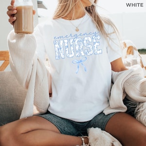 Puede incluir: Camiseta blanca con un dise&ntilde;o floral azul y el texto "emergency nurse" con un lazo azul.
