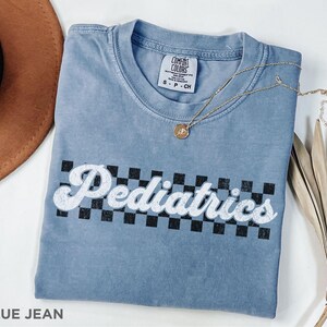 May include: A blue jean t-shirt with a white graphic that says "Pediatrics" in a retro font with a black and white checkered background.