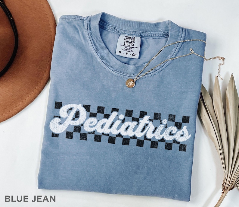 May include: A blue jean t-shirt with a white graphic that says "Pediatrics" in a retro font with a black and white checkered background.