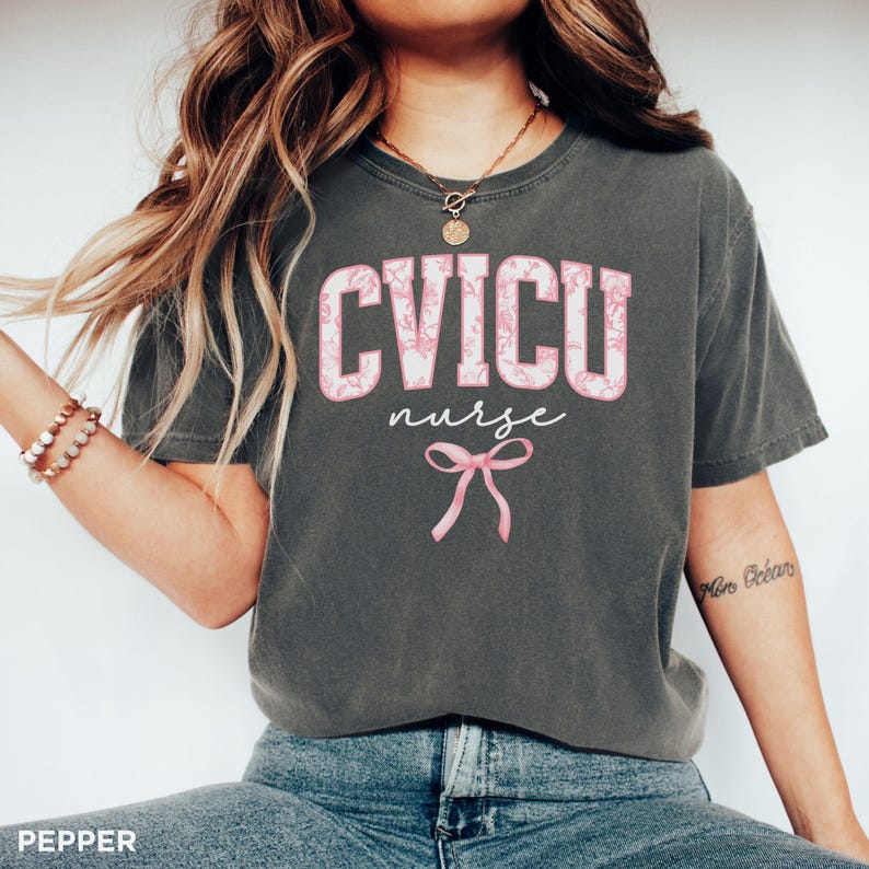 May include: A dark gray t-shirt with the text "CVICU nurse" in pink with a pink bow underneath. The shirt has a floral pattern on the text.