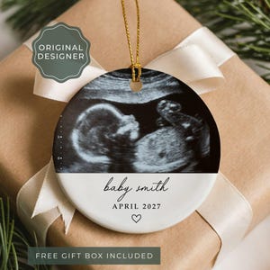 May include: A round white ceramic ornament featuring a black and white ultrasound image of a fetus at the top. Below the image, the text reads baby smith and APRIL 2027 with a small heart icon. A gold string is attached for hanging.