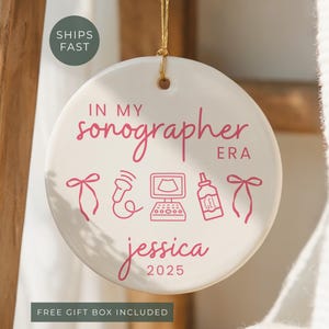 May include: A round, white ceramic ornament with a gold string for hanging. The ornament features pink text and line drawings related to sonography, including the words "IN MY sonographer ERA" and the name "jessica 2025".