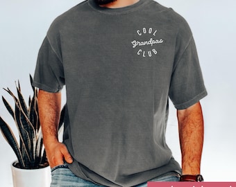 May include: Pepper-colored, short-sleeved t-shirt with the text "Cool Grandpas Club" in a white, circular design on the upper left chest. The shirt is embroidered and has a relaxed fit.