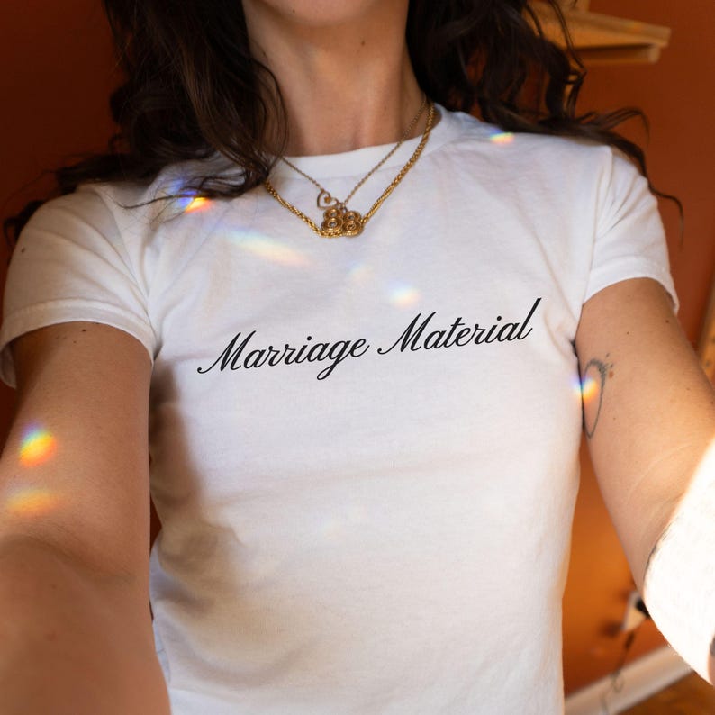 May include: White t-shirt with the phrase "Marriage Material" in black script. The shirt has short sleeves and a crew neck. A gold necklace is visible around the neck.