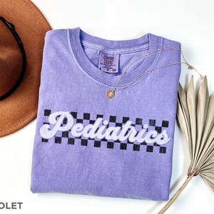 May include: A purple t-shirt with a white checkered pattern and the text "Pediatrics" in a retro font. The shirt is made by Comfort Colors.