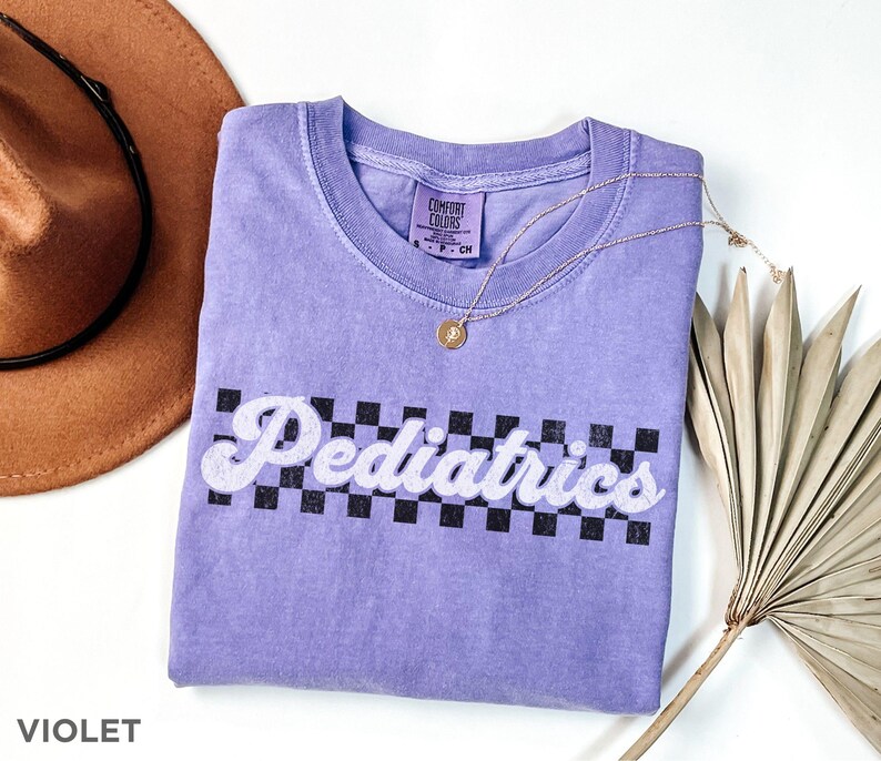 May include: A purple t-shirt with a white checkered pattern and the text "Pediatrics" in a retro font. The shirt is made by Comfort Colors.