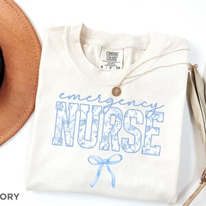 Puede incluir: Camiseta de color marfil con un dise&ntilde;o de estampado floral azul que dice "emergency nurse" con un lazo azul debajo.