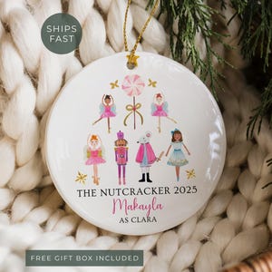 May include: White ceramic Christmas ornament featuring a Nutcracker ballet theme. The ornament has illustrations of ballerinas, a nutcracker, and a mouse. Text includes "The Nutcracker 2025" and a name. Gold string for hanging.