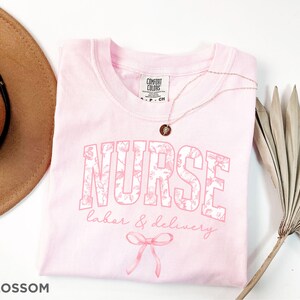 May include: Pink t-shirt with the word "NURSE" in white with floral print and the text "labor & delivery" in pink script. The shirt has a bow at the bottom.