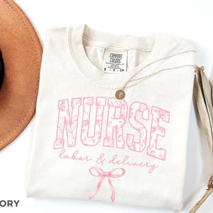 May include: Ivory t-shirt with a pink floral design that says "NURSE labor & delivery" with a pink bow underneath. The shirt has a tag that says "COMFORT COLORS".