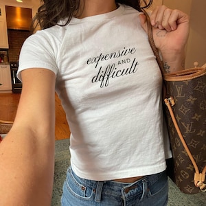 May include: White t-shirt with the text "expensive and difficult" in black script. The shirt is paired with blue jeans. A brown handbag with a monogram pattern is visible.