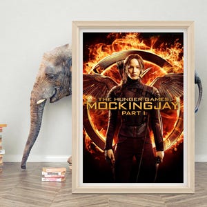 May include: A framed movie poster for "The Hunger Games: Mockingjay - Part 1." The poster shows a woman in black with wings, set against a fiery background. The poster is in a light wood frame.