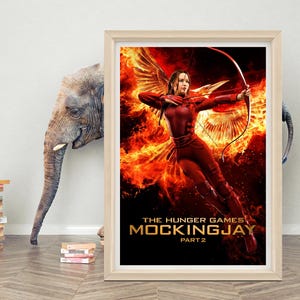 May include: Framed poster for "The Hunger Games: Mockingjay - Part 2." The image shows a woman in a red outfit with a bow and arrow, set against a fiery background. The poster is in a light-coloured wooden frame.