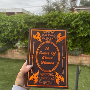 May include: A Court of Silver Flames book with a brown cover and orange flame and decorative accents. The title is in orange lettering. The book is held in front of a green background.