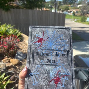 May include: A book titled "Web of Silence Duet" by Leigh Rivers. The cover features a gray background with a spiderweb design in light blue and red spiders. The book is held outdoors.