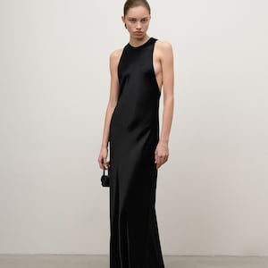 Puede incluir: Un vestido largo negro de satén con cuello alto y laterales abiertos. El vestido tiene un diseño sencillo y elegante, y es adecuado para eventos formales. Se sostiene un pequeño bolso negro en la mano.