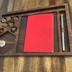 May include: A wooden tray with a red notebook, a pen, and a dice rolling area. The tray has a hexagonal cutout for a miniature figure.