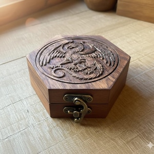 Puede incluir: Caja hexagonal de madera con un diseño de águila tallada en la tapa. La caja es de madera de tonos cálidos y tiene un cierre metálico. El tallado del águila es detallado, agregando un elemento decorativo a la caja.