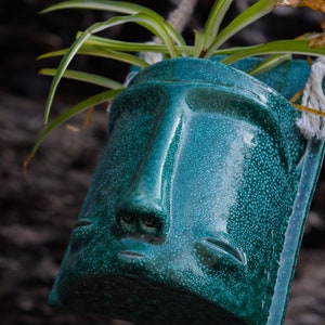 May include: A teal green ceramic planter shaped like a face with a plant growing out of the top. The planter has a textured surface and a glossy finish.