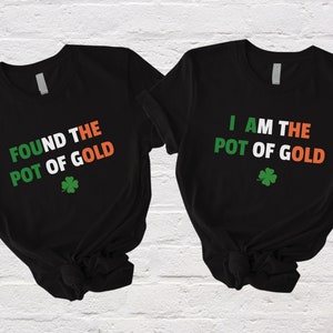 Könnte beinhalten: Zwei schwarze T-Shirts mit einem weißen, orangen und grünen Design. Ein Shirt sagt "FOUND THE POT OF GOLD" und das andere sagt "I AM THE POT OF GOLD". Beide Shirts haben ein grünes Kleeblatt.