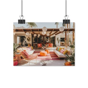 Op de afbeelding: Een buitenpatio met een mid-century modern design met een witte bank, twee oranje stoelen en een roze en oranje gestreept tapijt. De patio is versierd met witte papieren lantaarns en weelderig groen.