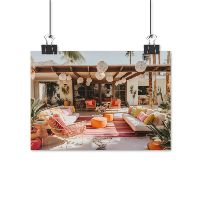 Op de afbeelding: Een buitenpatio met een mid-century modern design met een witte bank, twee oranje stoelen en een roze en oranje gestreept tapijt. De patio is versierd met witte papieren lantaarns en weelderig groen.