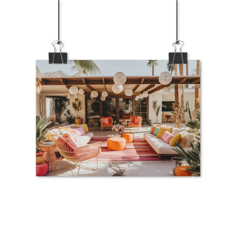 Op de afbeelding: Een buitenpatio met een mid-century modern design met een witte bank, twee oranje stoelen en een roze en oranje gestreept tapijt. De patio is versierd met witte papieren lantaarns en weelderig groen.