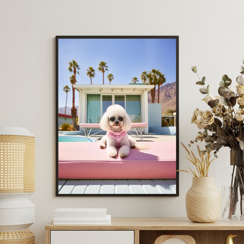 K&ouml;nnte beinhalten: Ein gerahmter Wandkunstdruck, der einen wei&szlig;en Pudel mit schwarzer Sonnenbrille und rosa Geschirr zeigt, der auf einer rosa Poolliege liegt. Im Hintergrund sind ein modernes Haus, Palmen, ein Swimmingpool und ein Gebirge zu sehen.
