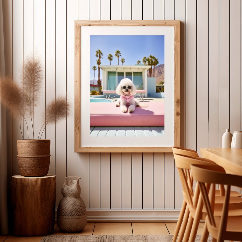 K&ouml;nnte beinhalten: Ein gerahmter Kunstdruck, der einen wei&szlig;en Pudel mit schwarzer Sonnenbrille und rosa Geschirr zeigt, der auf einer rosa Pool-Liege vor einem modernen Haus mit Palmen unter einem klaren blauen Himmel ruht, in einem hellen Holzrahmen.