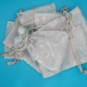 May include: A pile of white organza gift bags with silver drawstrings. The bags are made of a sheer, delicate fabric and are ideal for small gifts or favours.