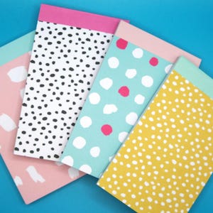 May include: Four different patterned notepads with colourful borders. The notepads have a polka dot pattern in black and white, pink and white, blue and white, and yellow and white.