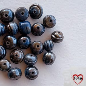 Könnte beinhalten: Eine Sammlung dunkelblauer und grauer Perlen mit metallischem Glanz. Die Perlen sind rund, einige mit gerippten Details, und haben ein Loch in der Mitte zum Auffädeln. Die Perlen sind auf einer weißen Oberfläche verstreut.